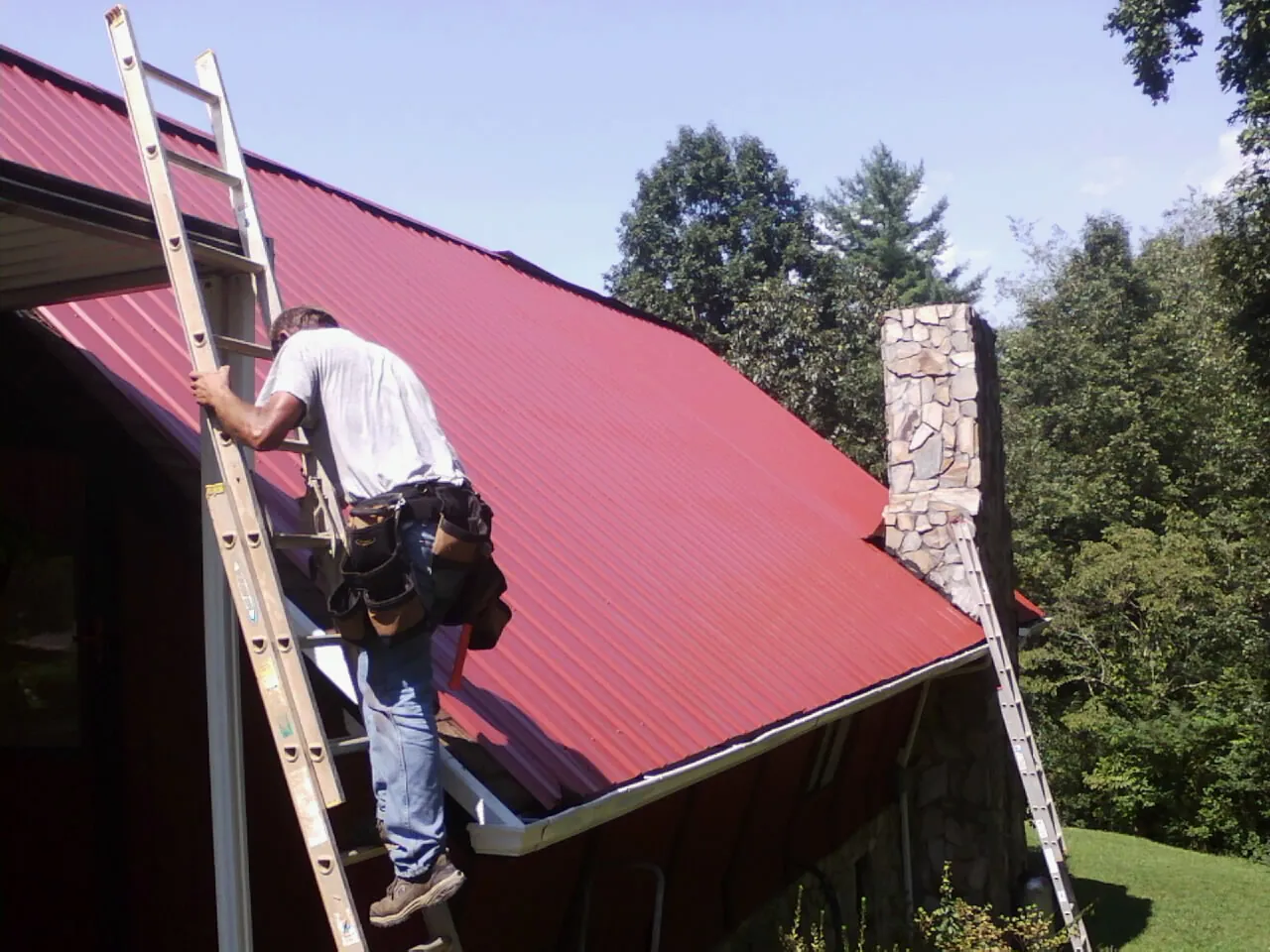 Roofer on a ladder beside a red metal roof and stone chimney — Soffit & Fascia Repair by Roofing Brothers in Marlton